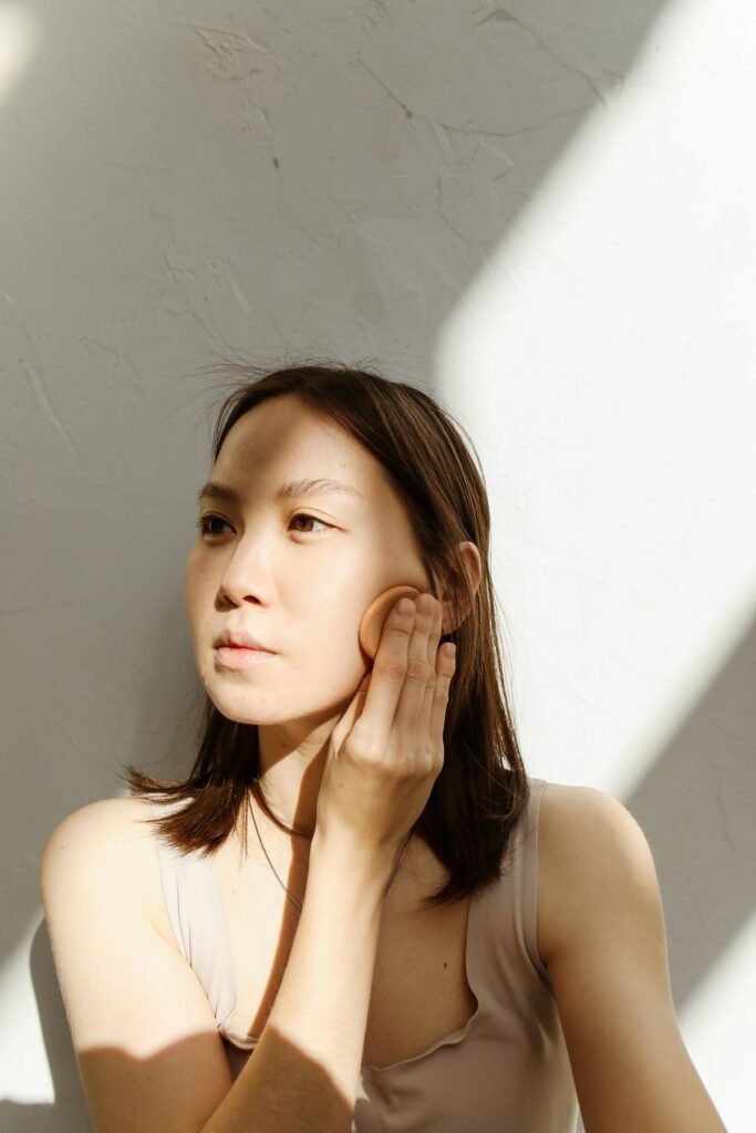 A woman in soft window light gently touching her cheek, skin natural and luminous