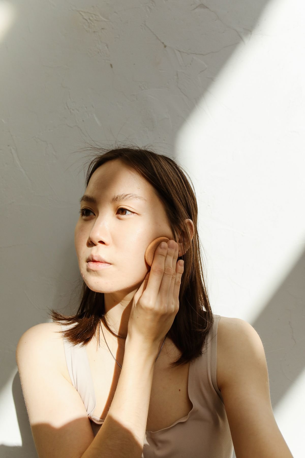 A woman in soft window light gently touching her cheek, skin natural and luminous